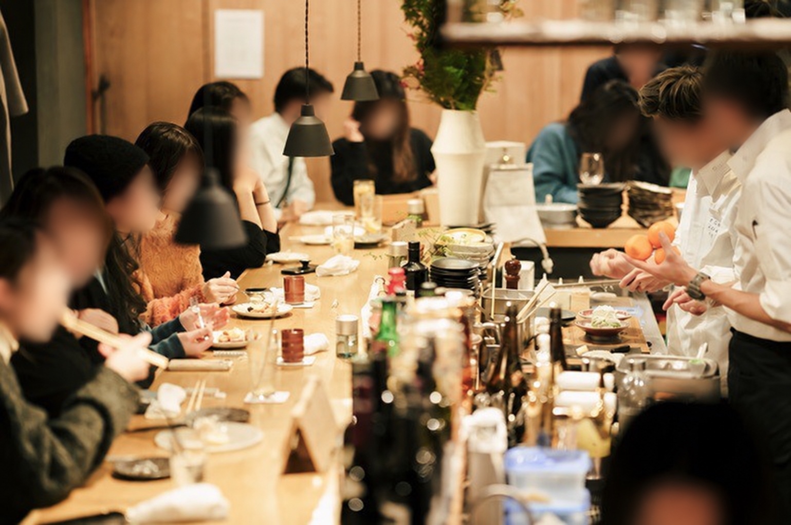 カウンター席に客が並び、手前にお酒の瓶とグラス、奥で料理人が盛り付けている